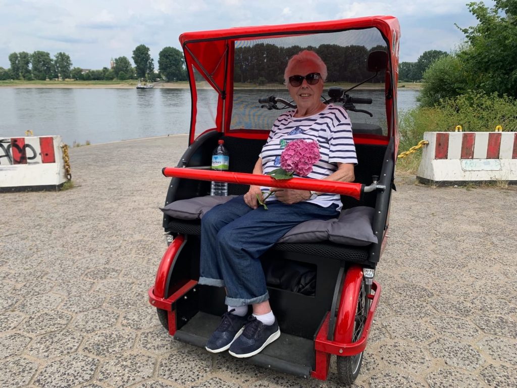 Seniorin sitzt in einer Rikscha am Rhein